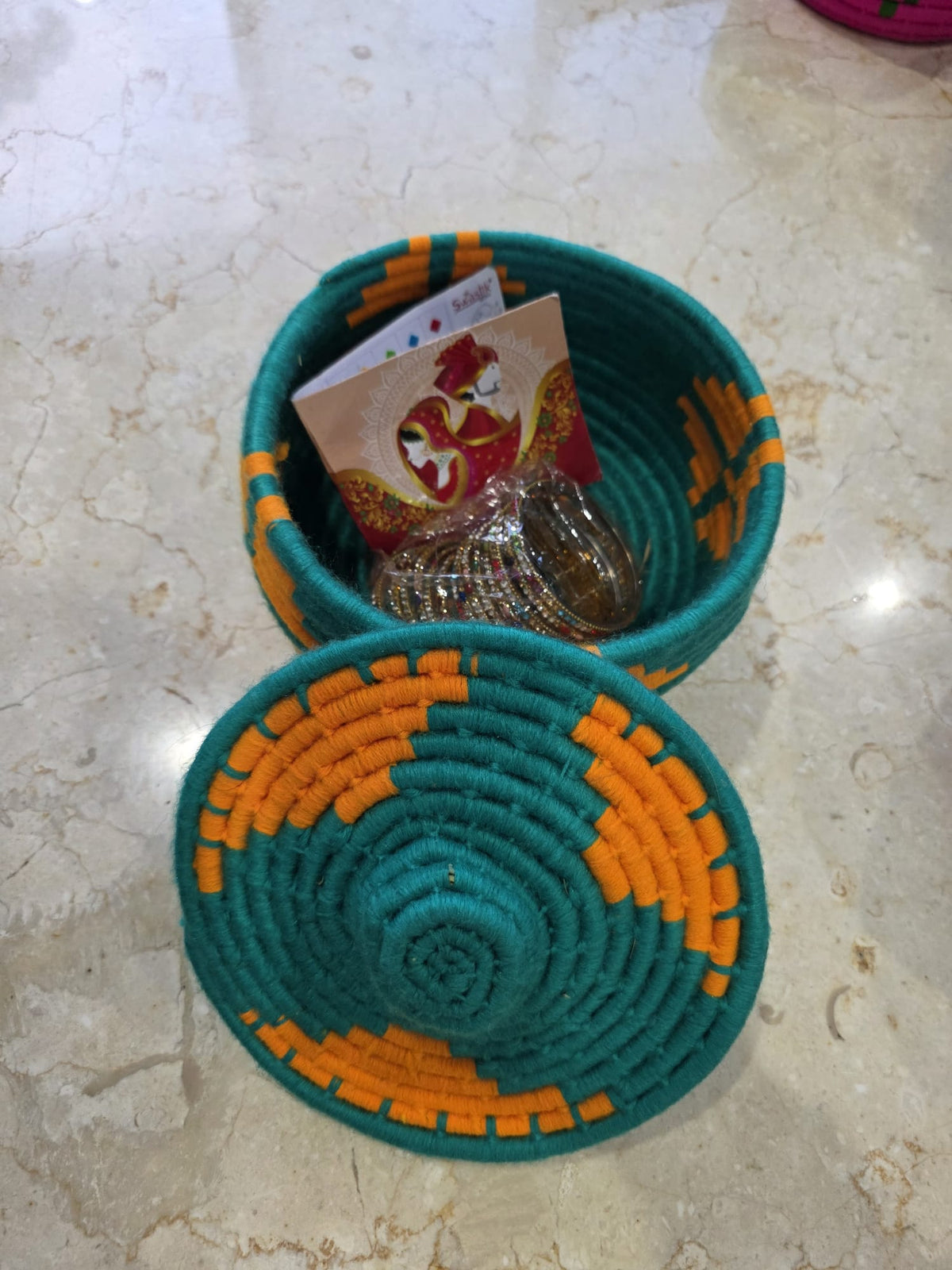 A teal and orange woven basket with its matching lid placed in front, containing a small gift box and a card.