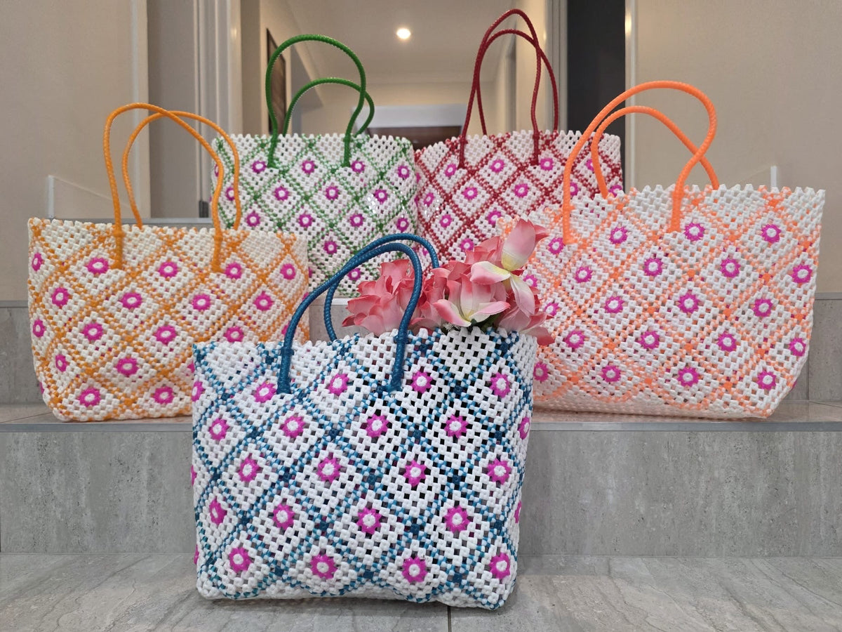 Four woven tote bags with colorful diamond and floral patterns are displayed on gray stone steps. The front bag is white with blue and pink patterns and blue handles, with pink flowers tucked inside.