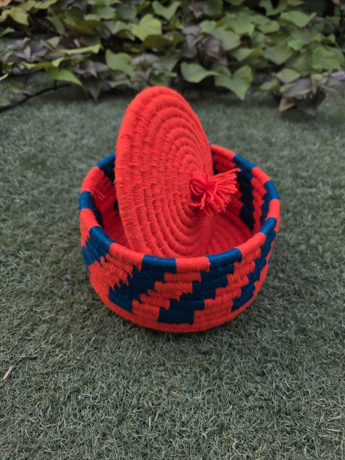 A small woven basket with a matching lid, featuring a red and dark blue geometric pattern.