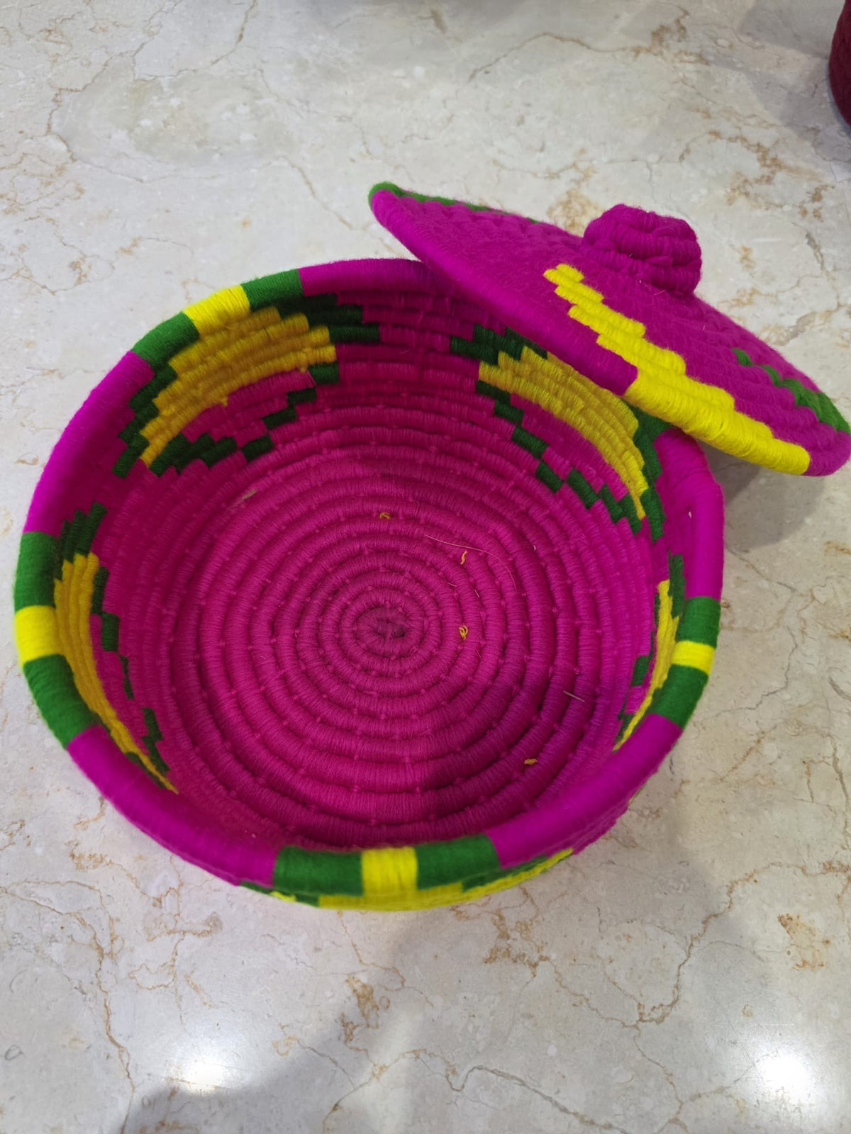 A hand-woven basket with a matching lid, featuring a bright magenta-pink body with yellow and green geometric patterns, placed on a light marble surface.